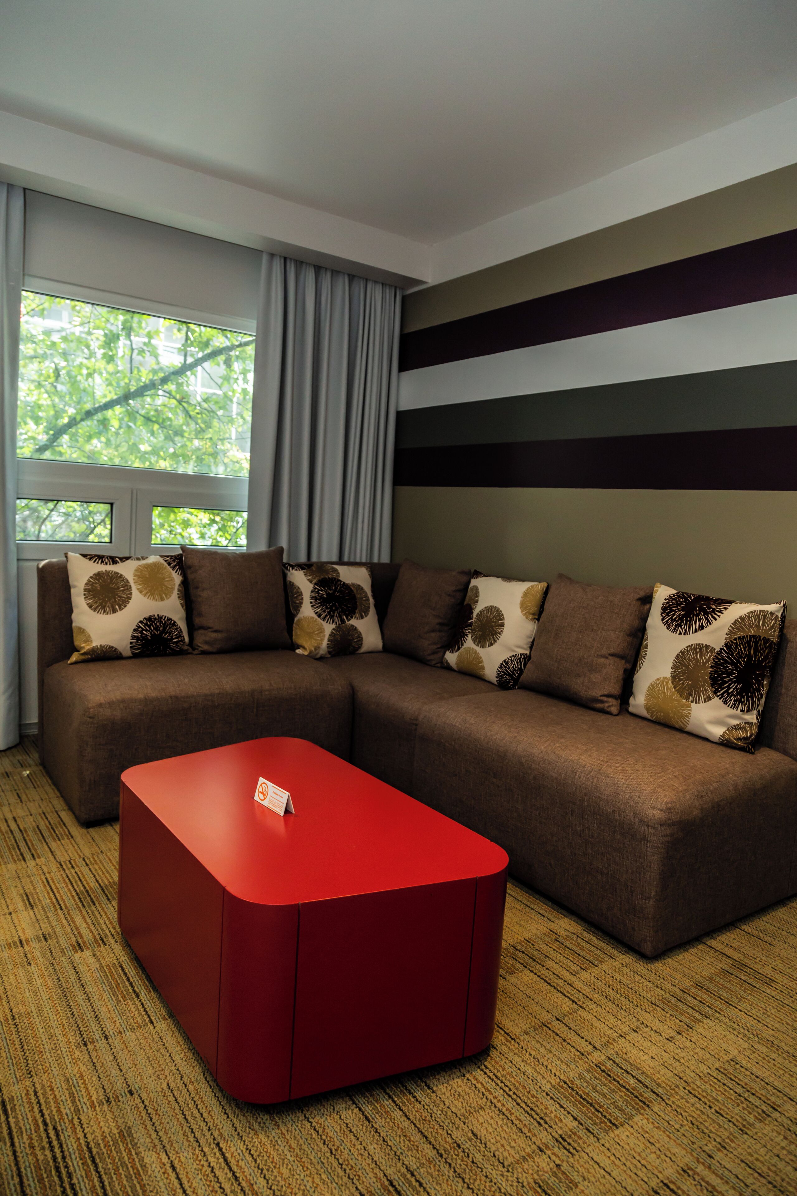 Una sala moderna con un sofá modular, un reposapiés rojo y una ventana con cortinas.