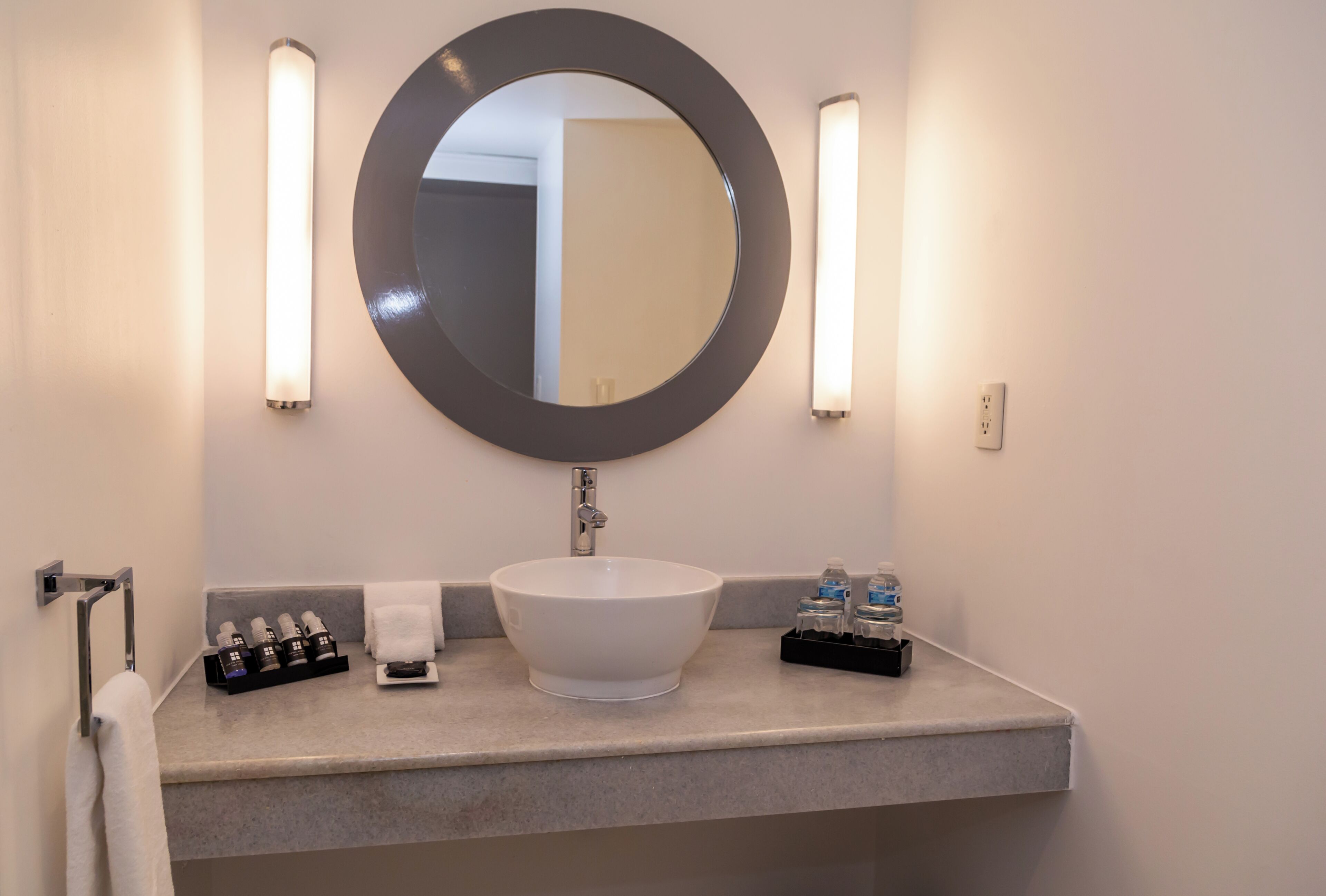 A bathroom with a round mirror, a white sink, two wall-mounted lights, a towel rack, and a small shelf with toiletries.