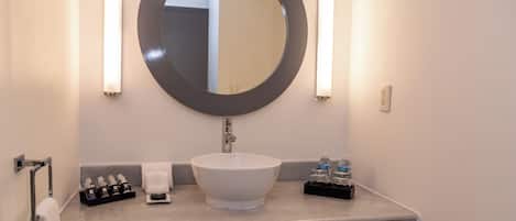 Un baño con un espejo redondo, un lavamanos blanco, dos luces empotradas en la pared, un perchero y un pequeño estante con artículos de tocador.