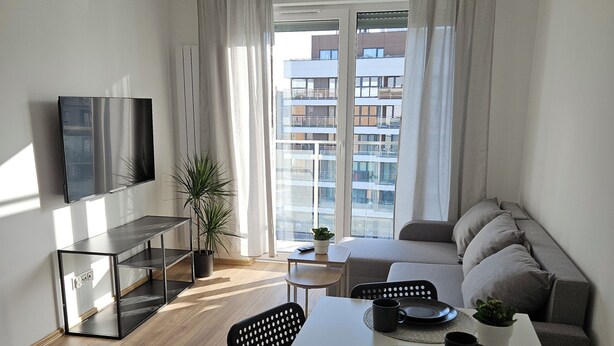 A modern living room with a dining area, a flat-screen TV mounted on the wall, a sofa, and a large window with curtains. - Malta View Prestige (Poznań)