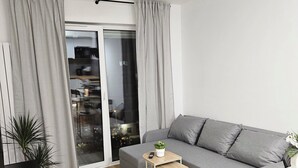 A modern living room with a grey sofa, a small wooden side table, a black side table, a white rug, and a potted plant. - Malta View Prestige (Poznań)