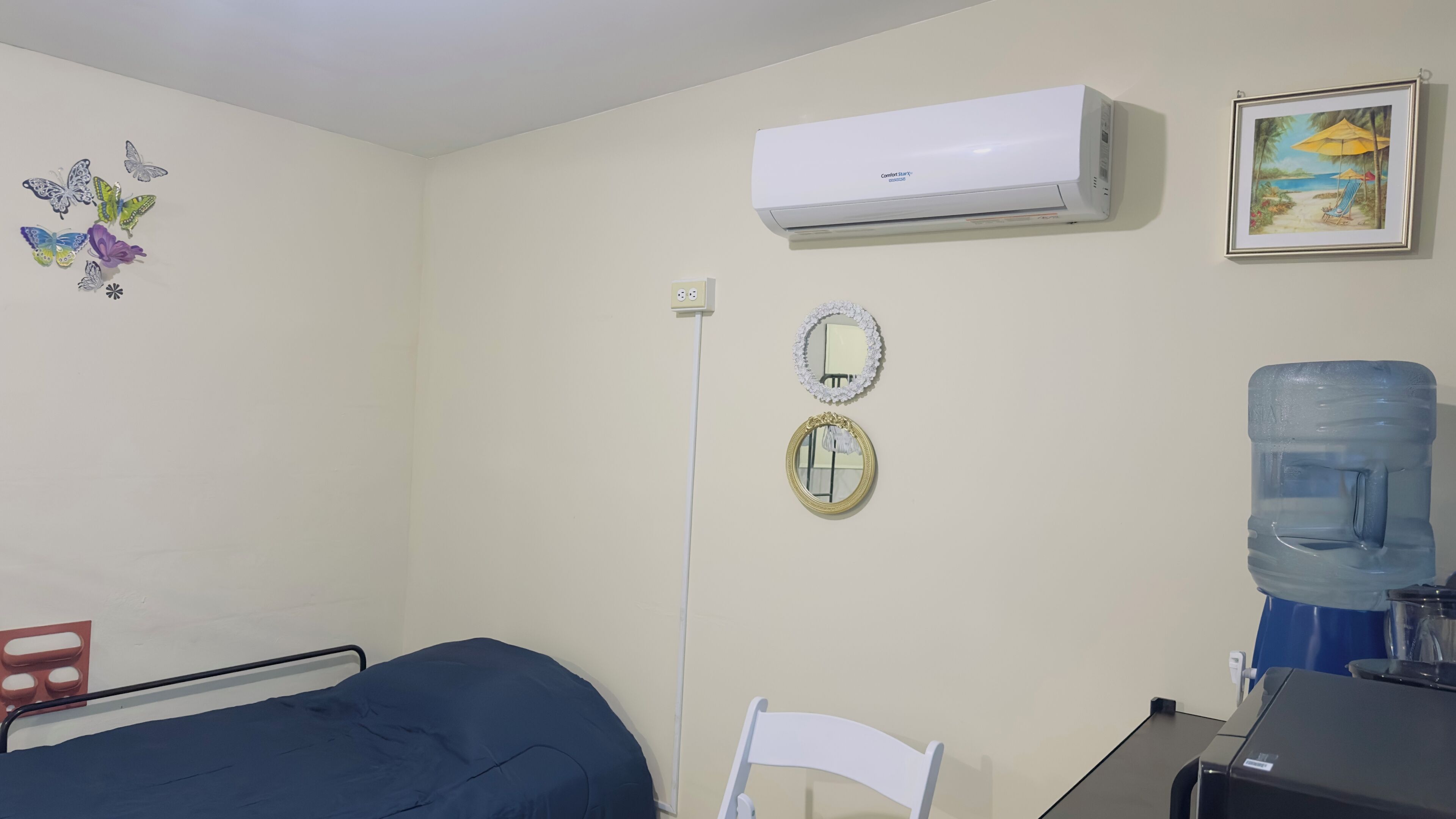 A bedroom with a bed, a chair, a desk, an air conditioner, a water dispenser, and decorative wall items.