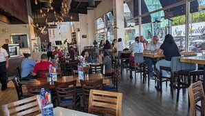 Un interior de restaurante con mesas y sillas de madera, lámparas colgantes y ventanas amplias.