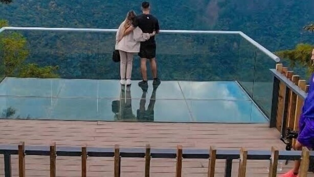 Una pareja parada sobre un piso de vidrio con vistas a un paisaje montañoso.