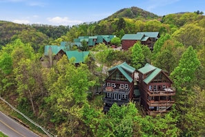 A cluster of log cabins with green roofs, surrounded by dense forest, and a road running alongside. - Ober Overlook: Views, Pool, Hot Tub & Game Area! (Gatlinburg)