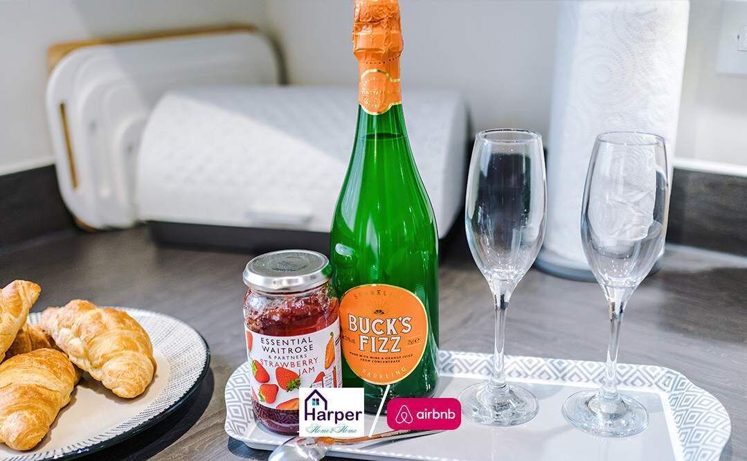 A breakfast setup with croissants, a bottle of Buck's Fizz, a jar of strawberry jam, and two wine glasses on a tray.