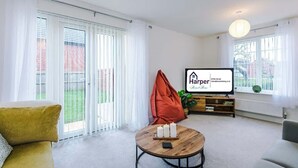 Ein modernes Wohnzimmer mit einer Couch, einem Couchtisch mit Kerzen, einem Fernseher und Blick auf einen Garten durch große Fenster.
