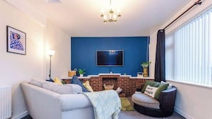 Ein Wohnzimmer mit Sofa, Sessel, Kamin, Fernseher und einem Fenster mit Jalousien.