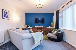Ein Wohnzimmer mit Sofa, Sessel, Kamin, Fernseher und einem Fenster mit Jalousien.