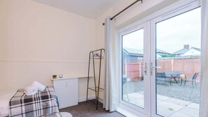 Ein kleines Zimmer mit einem Bett, einem Metallregal und einer Schiebetür zu einem Außenbereich mit einer Bank und einem Tisch.