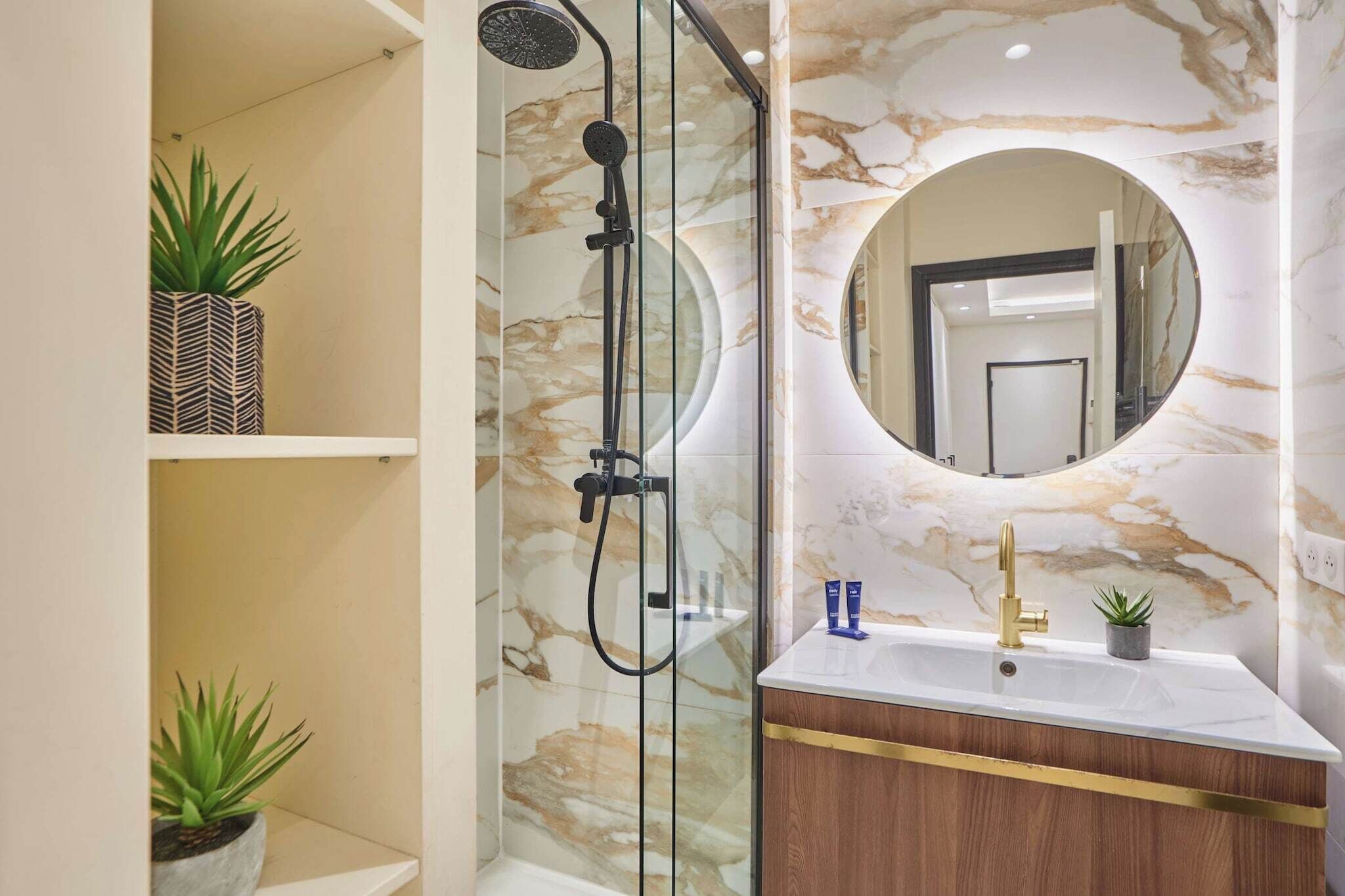 Une salle de bain dotée d’une douche à parois de verre, d’un mur en marbre, d’un miroir rond et d’un meuble-lavabo en bois avec des détails dorés.