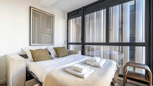 Een slaapkamer met een bed, kussens, een groot raam en een stoel.