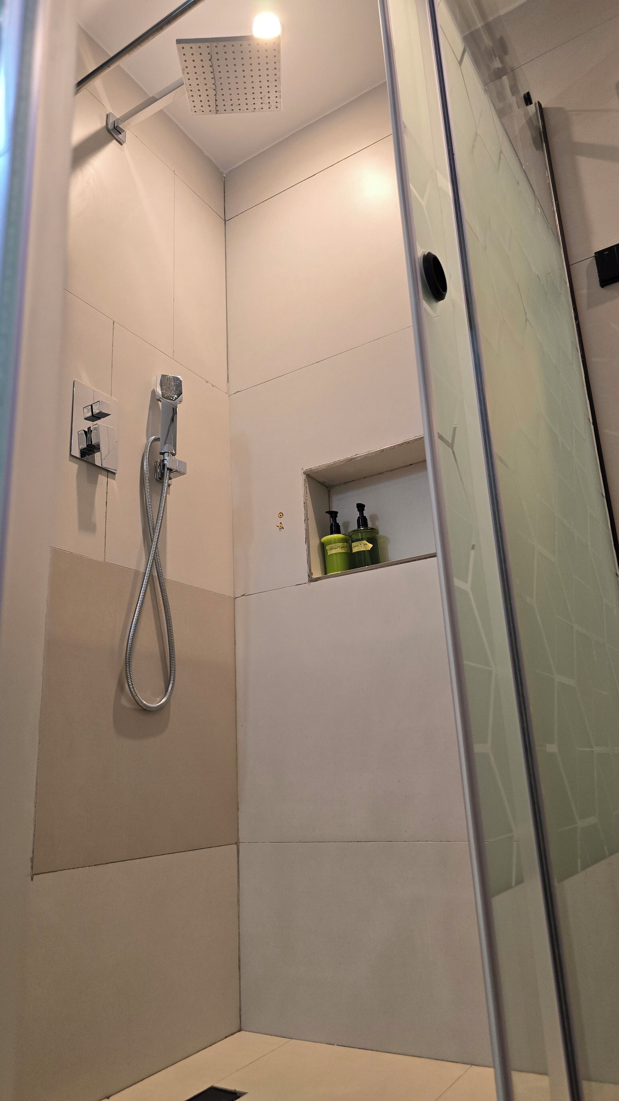 Chuveiro com chuveiro manual, área de chuveiro com divisória de vidro e prateleira com produtos de higiene.