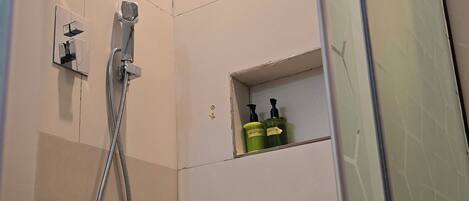 Chuveiro com chuveiro manual, área de chuveiro com divisória de vidro e prateleira com produtos de higiene.