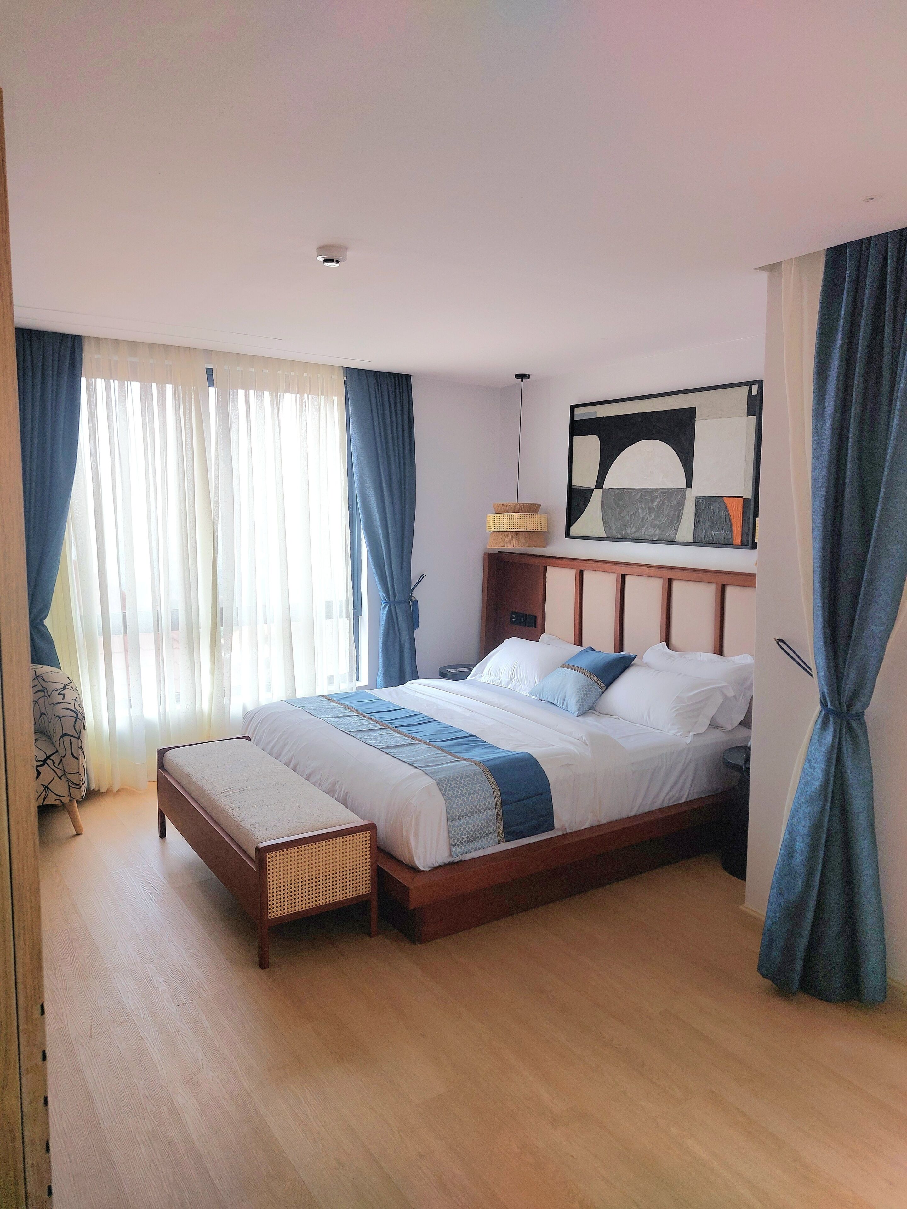 A modern hotel room with a wooden bed, a bedside table, a lamp, and a large abstract painting on the wall.