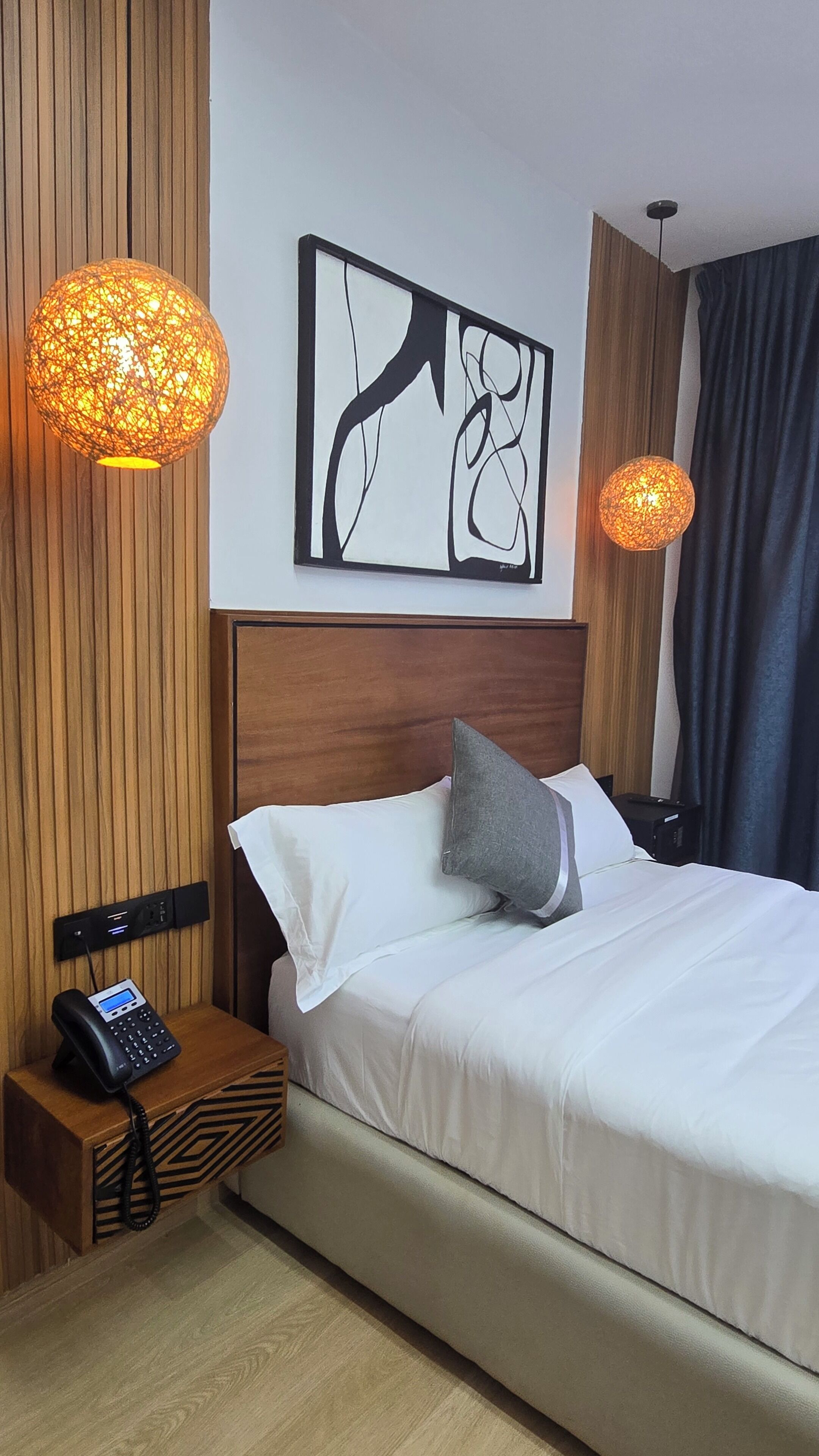 A hotel room with a bed, a bedside phone, a wall art piece, and two pendant lights.