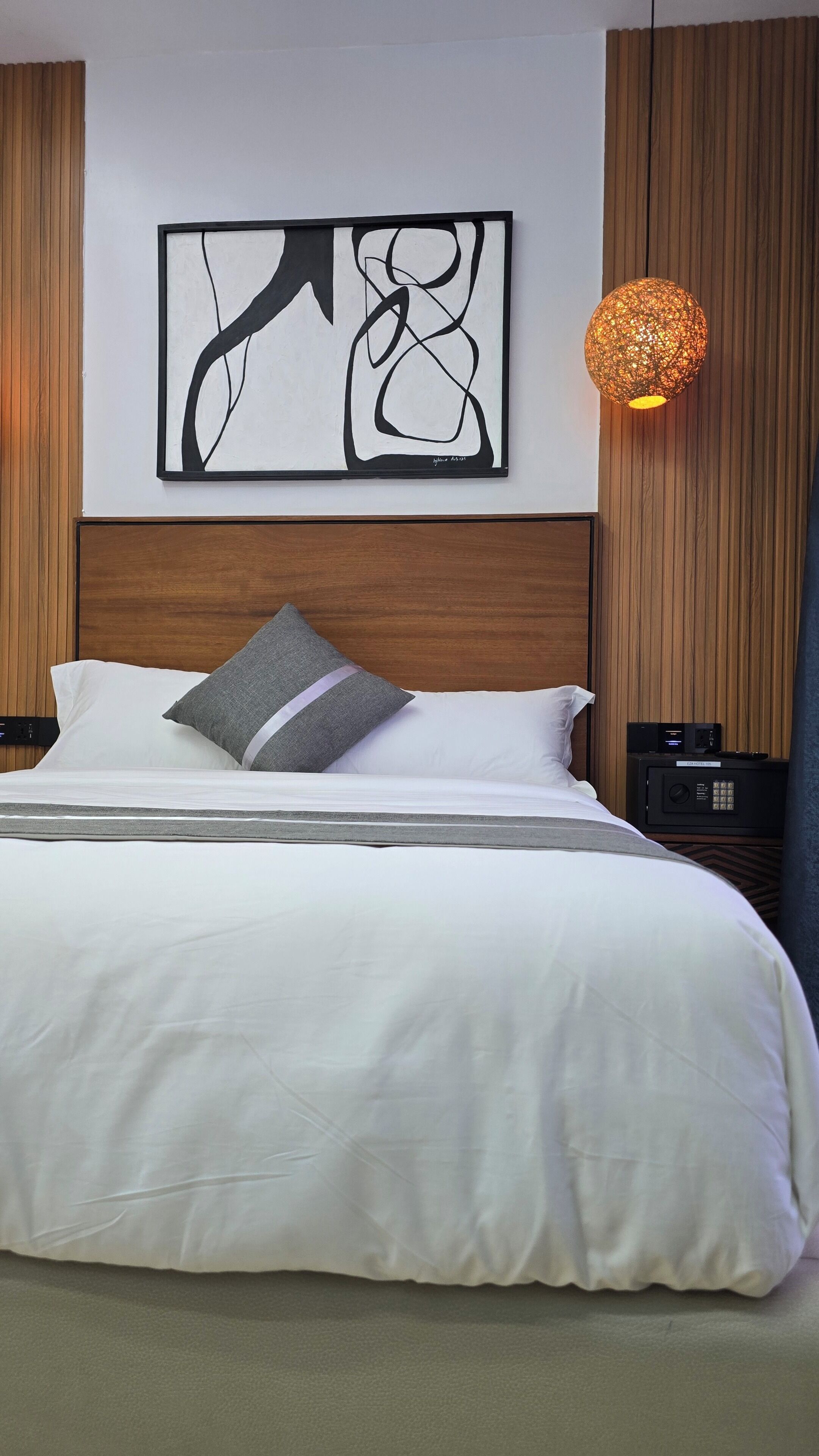 Habitación de hotel con una cama grande, un cuadro abstracto moderno, cabecera de madera, mesita de noche con lámpara y panel de control.
