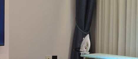A room with a wooden table, an ironing board, a wall-mounted air conditioner, and a curtain.