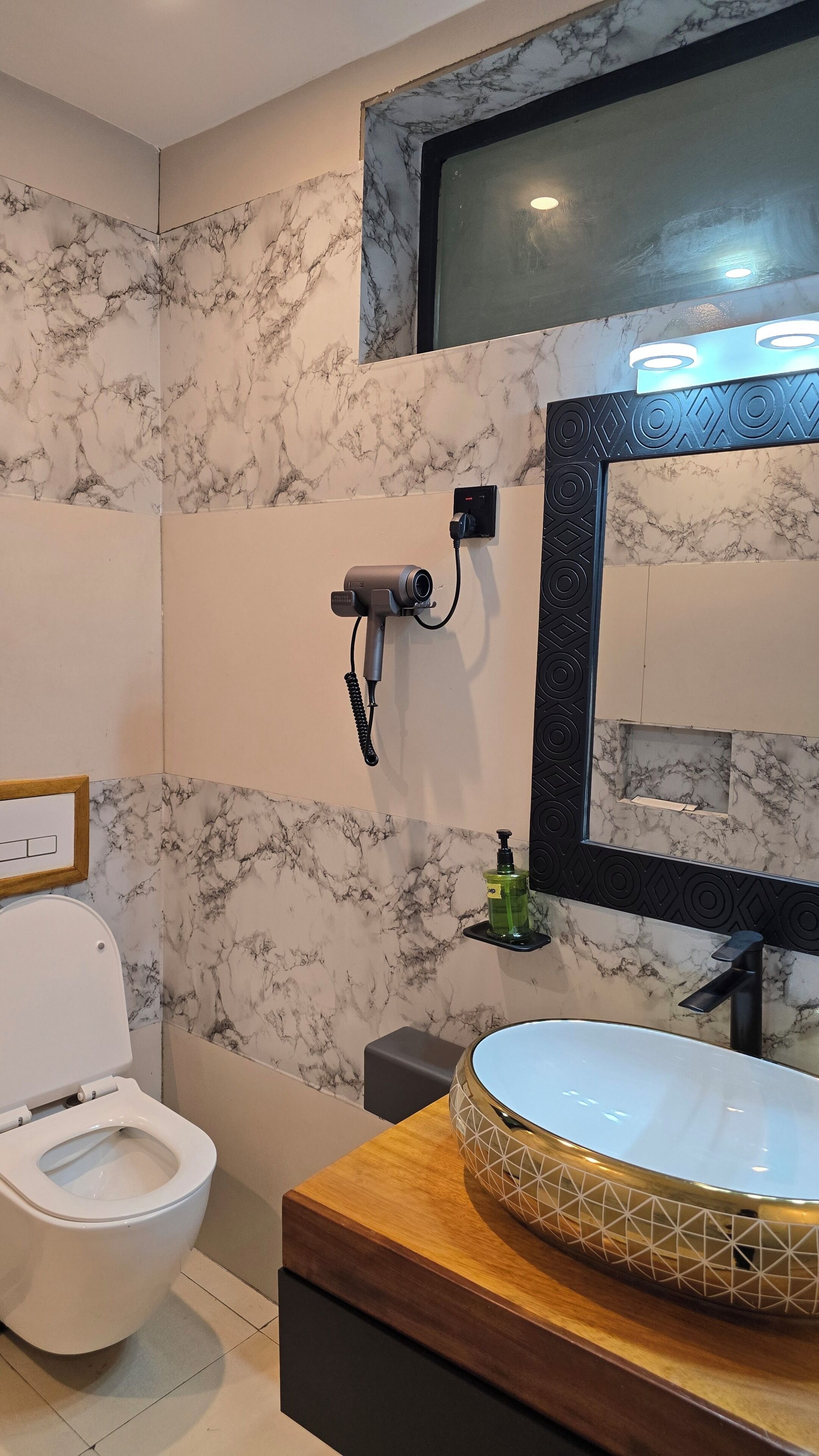 Un baño con inodoro, lavabo de mármol y secador de pelo en un soporte de pared, además de un espejo con marco negro.