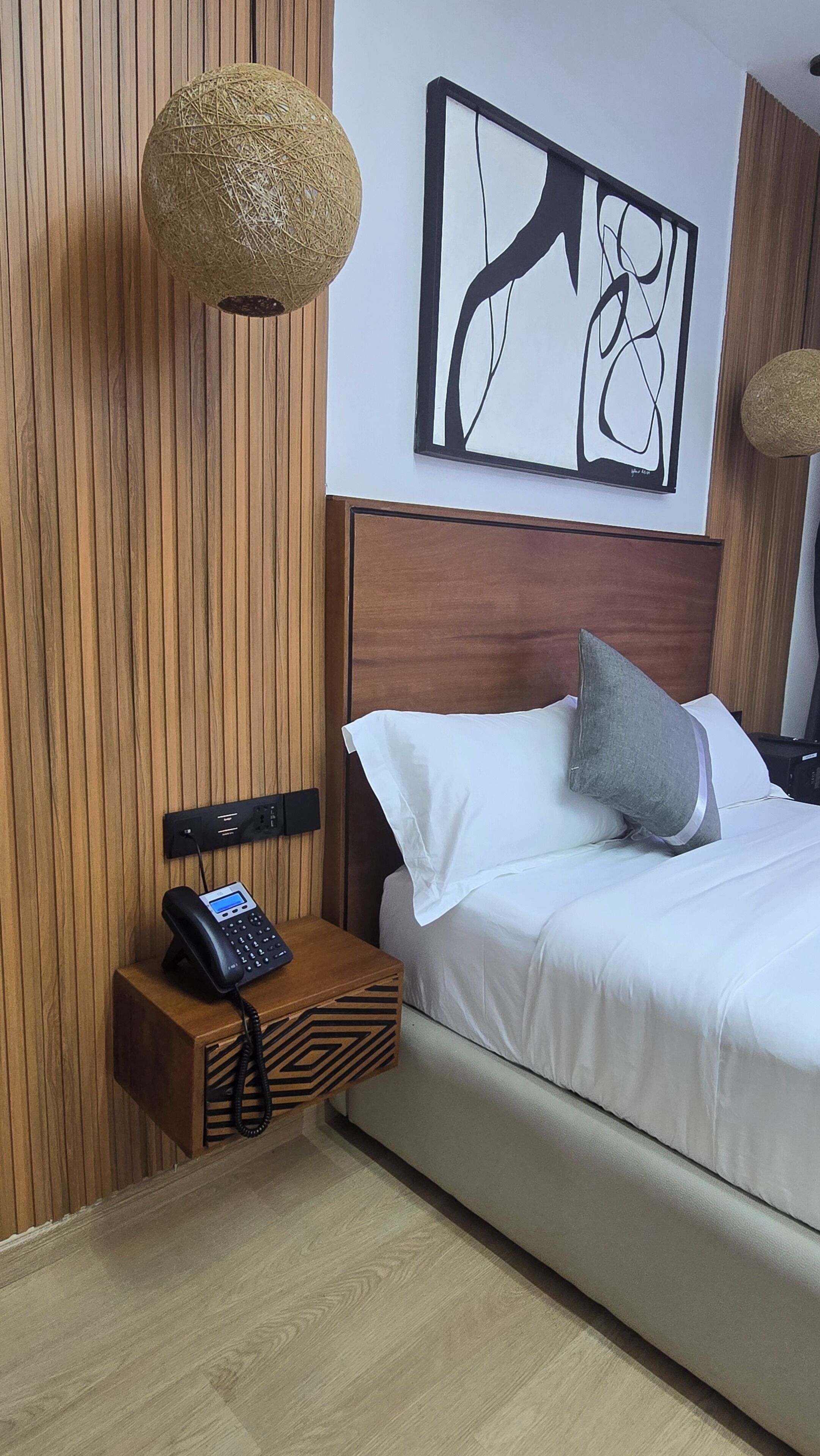 A hotel room with a wooden headboard, a bed with white linens, a gray pillow, a wall-mounted abstract art piece, a wooden bedside table with a telephone, and a textured spherical light fixture.