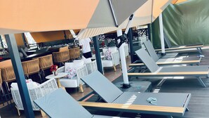 A sun lounge area with lounge chairs and tables, a covered seating area with wicker furniture, and a green tent-like structure in the background. - Eza Hotel (Kigali)
