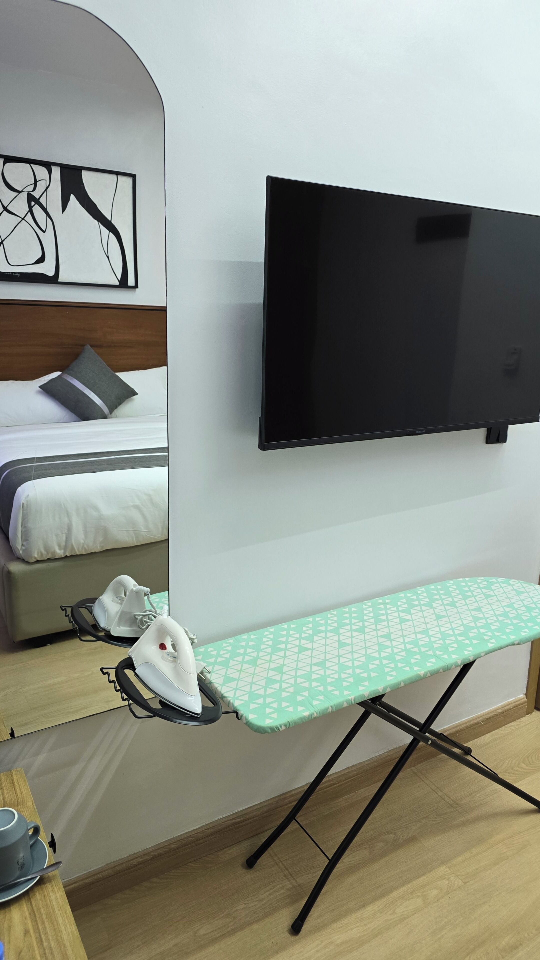 A hotel room with a bed, a flat-screen TV mounted on the wall, an ironing board with an iron, and a cup on a table.