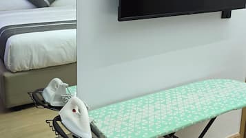 A hotel room with a bed, a flat-screen TV mounted on the wall, an ironing board with an iron, and a cup on a table.