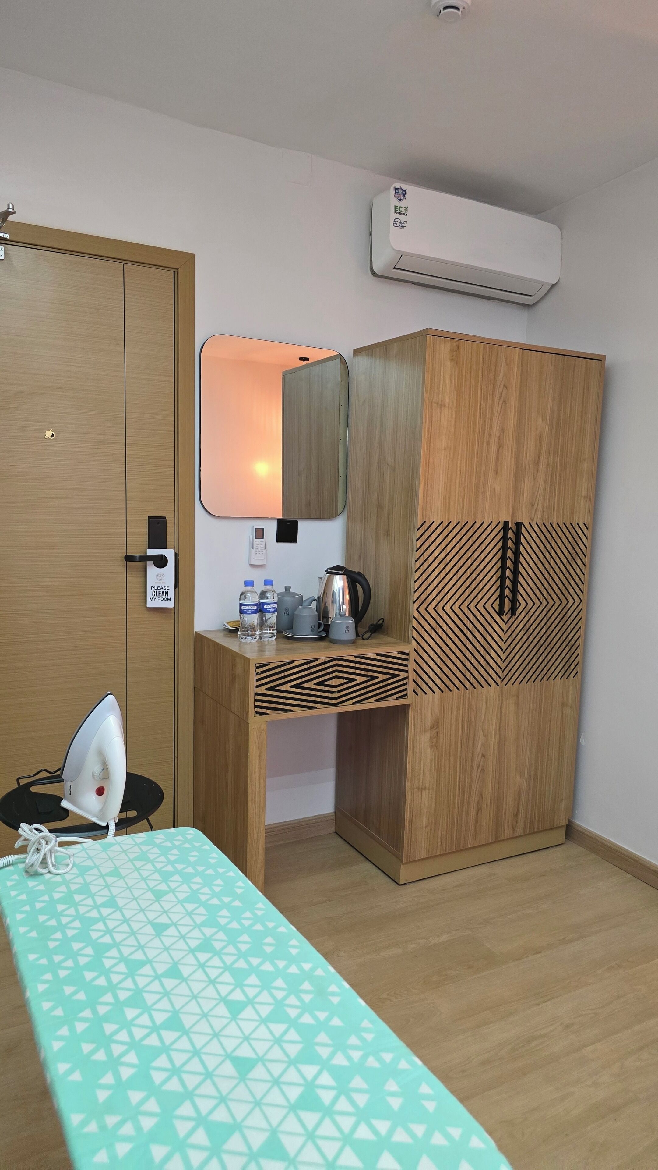 A room with a wooden wardrobe, a desk with a mirror, an ironing board, and a wall-mounted air conditioner.