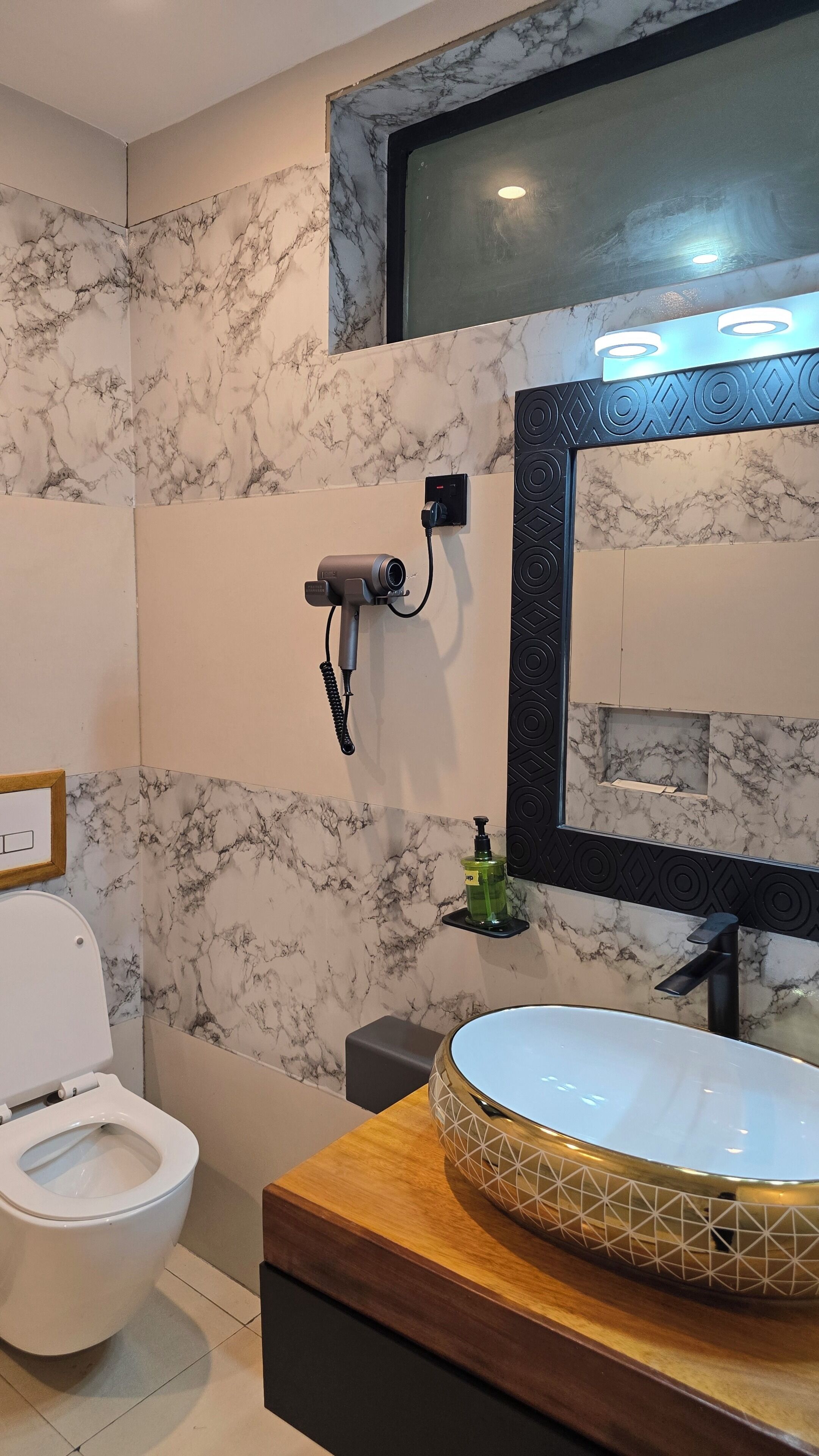 A bathroom with a marble wall, a toilet, a sink with a geometric pattern, and a hairdryer mounted on the wall.