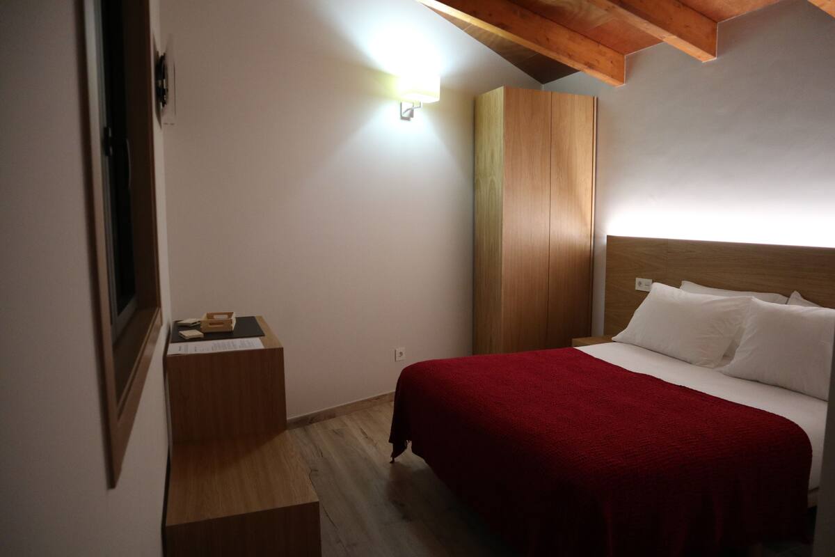 A bedroom with a wooden headboard, a red bedspread, a nightstand with a lamp, and a wooden floor.