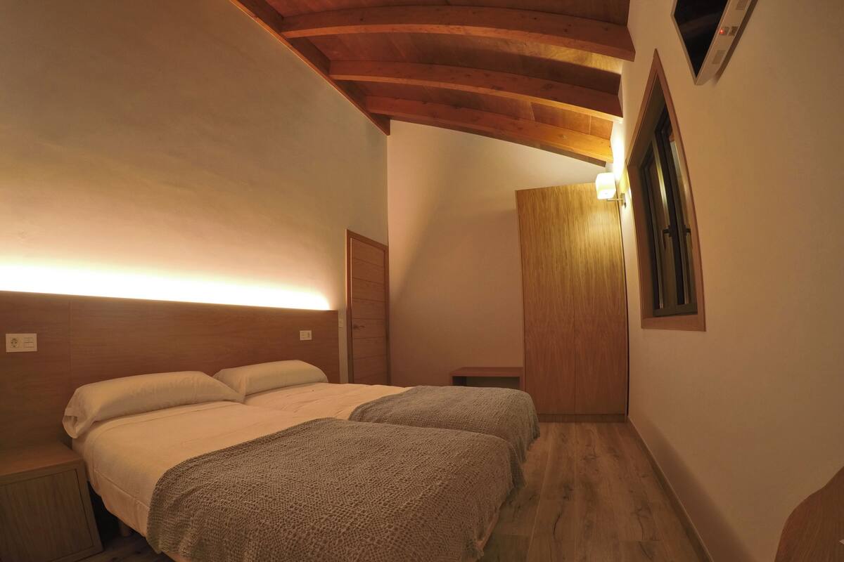 A bedroom with two beds, wooden beams on the ceiling, a small bedside table, and a window with a view.
