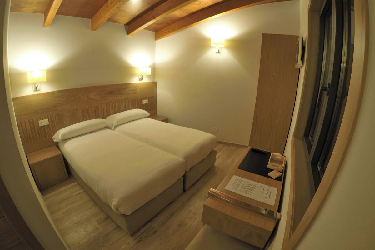 A room with a wooden ceiling, a bed with two pillows, a nightstand, a desk with a lamp, and a window.
