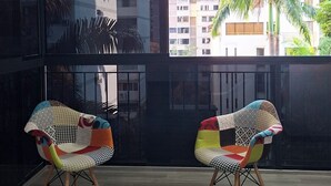 Une pièce avec deux chaises colorées, une grande fenêtre donnant sur des immeubles et des palmiers, et un plancher en bois.