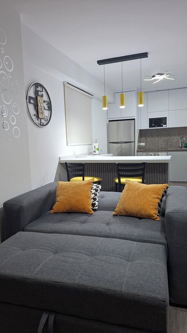A modern living room with a grey sofa, two mustard pillows, a wall clock, and a kitchen area with a microwave and a refrigerator.