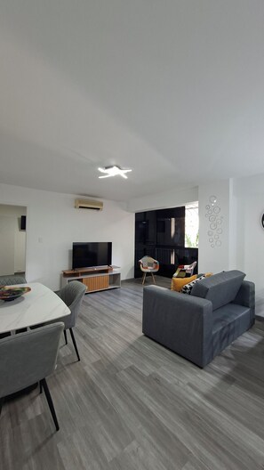 A modern living room with a flat-screen TV, grey sofa, dining table, and chairs.