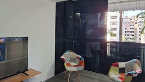 Un salon moderne avec un téléviseur à écran plat posé sur un meuble en bois, deux fauteuils colorés et une grande fenêtre donnant sur des immeubles.