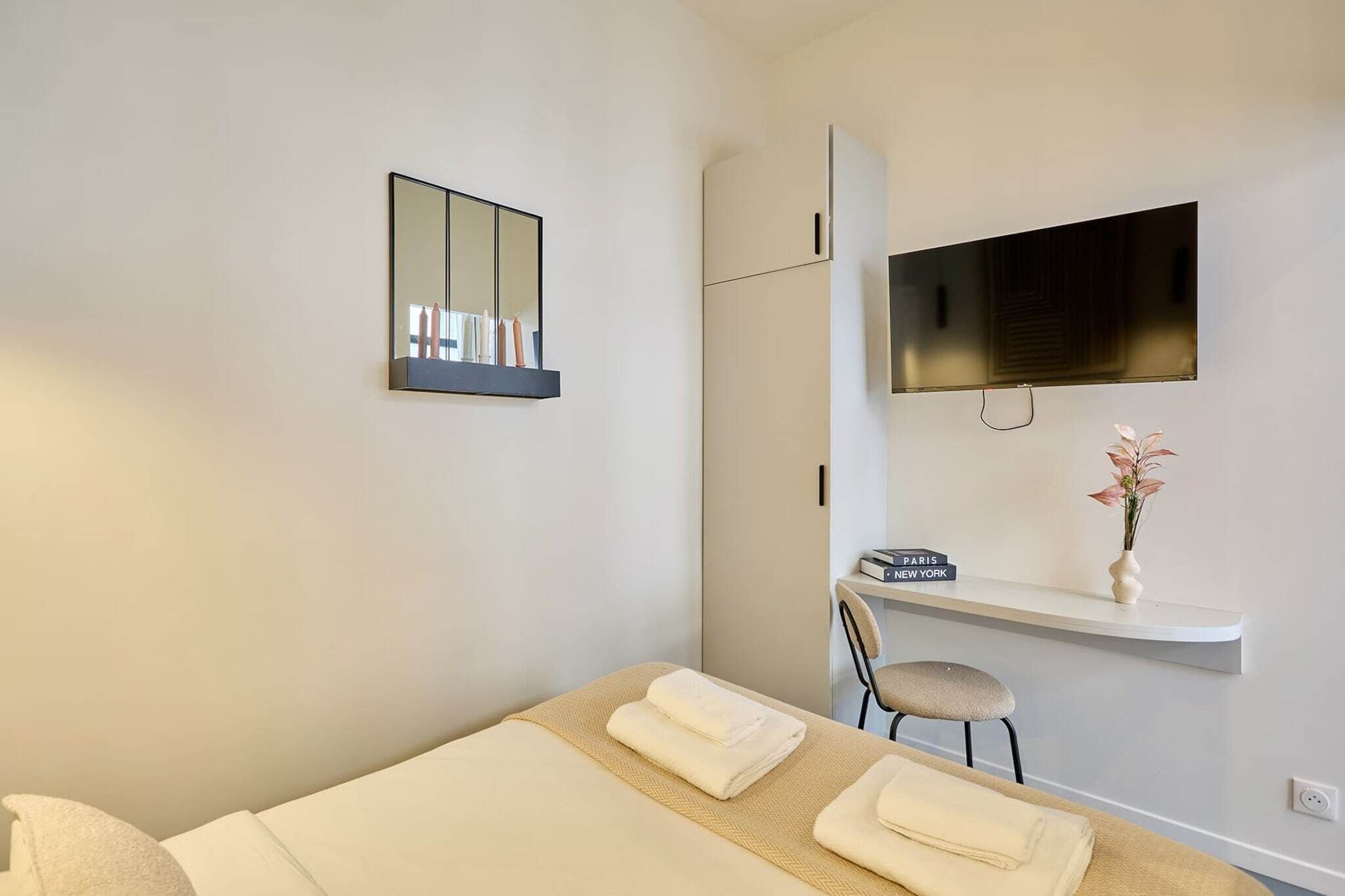 Una camera d'albergo con un letto, una poltrona, una TV, uno specchio e uno scaffale con un vaso e un cartello.
