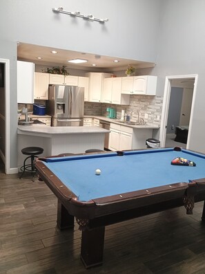 A pool table in a room with a kitchen area, including a refrigerator and cabinets.