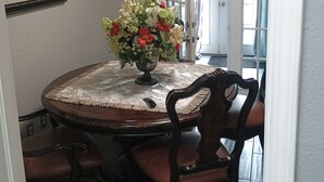 Et spiseområde med et rundt bord, stole, en blomsterdekoration i midten, et indrammet billede på væggen og en dør, der fører ud.