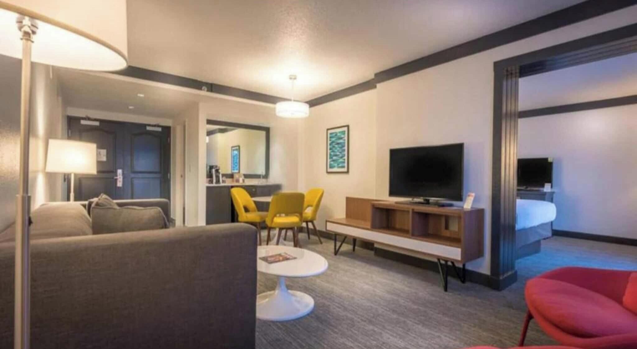 Habitación de hotel con sofá, mesa de comedor, televisión y una silla roja.