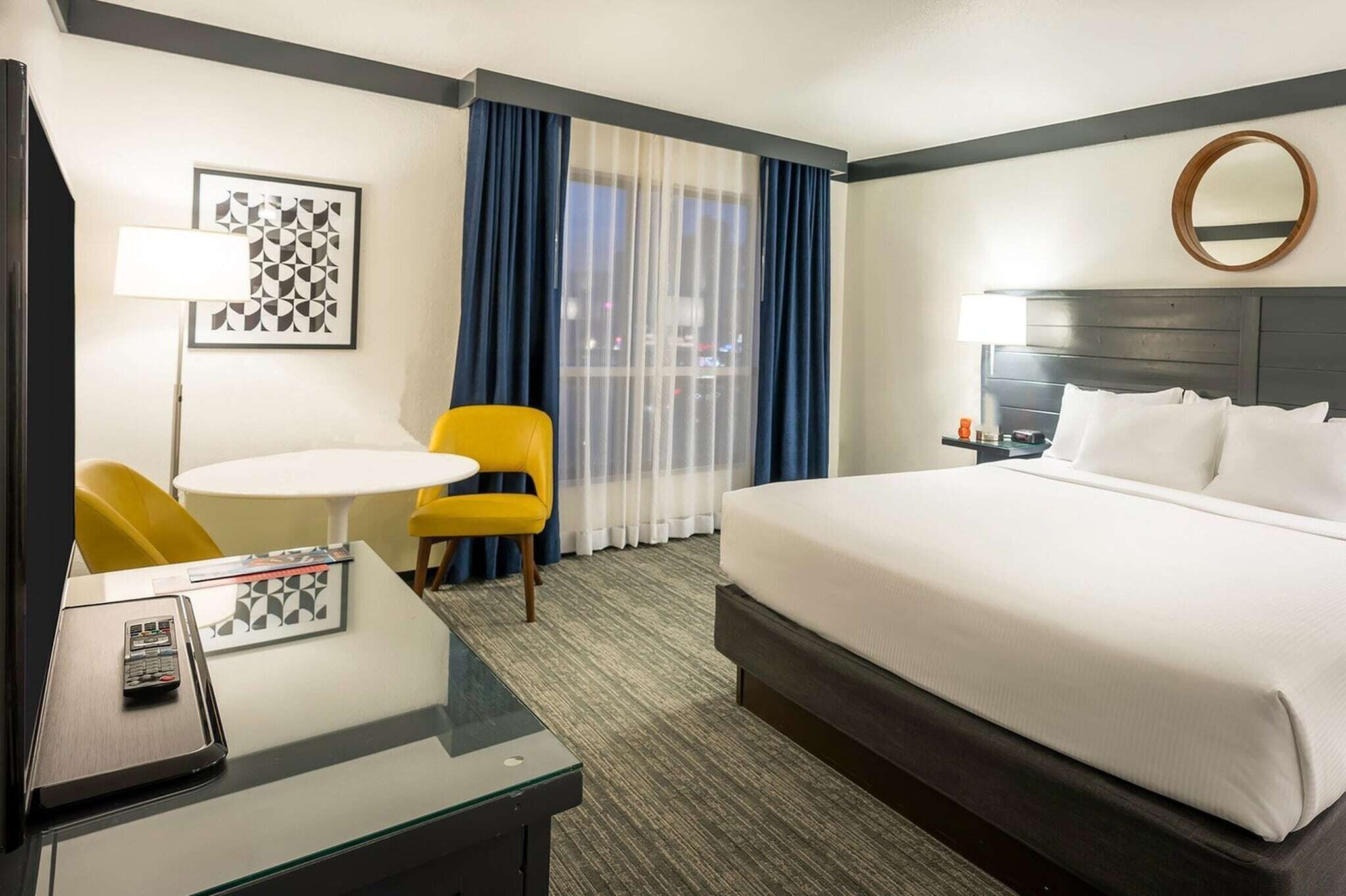 Una habitación de hotel con una cama, un escritorio con silla, una mesita, una lámpara, un cuadro enmarco y una ventana con cortinas.