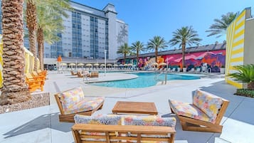 A poolside area with lounge chairs, a pool, palm trees, and a building with a mural.
