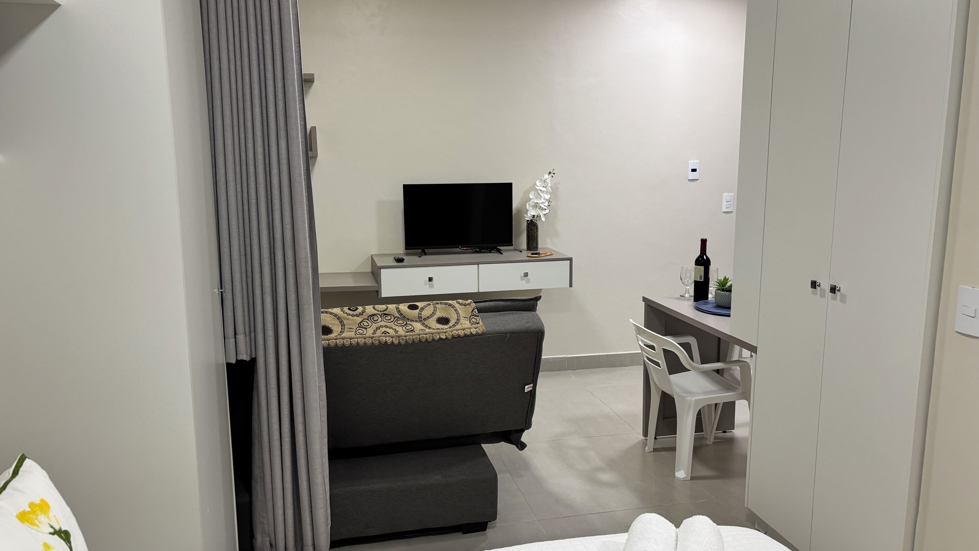 Um quarto moderno com um sofá cinzento, uma TV sobre um console branco, uma pequena mesa de trabalho com uma cadeira e um guarda-roupa.