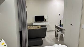 Um quarto moderno com um sofá cinzento, uma TV sobre um console branco, uma pequena mesa de trabalho com uma cadeira e um guarda-roupa.