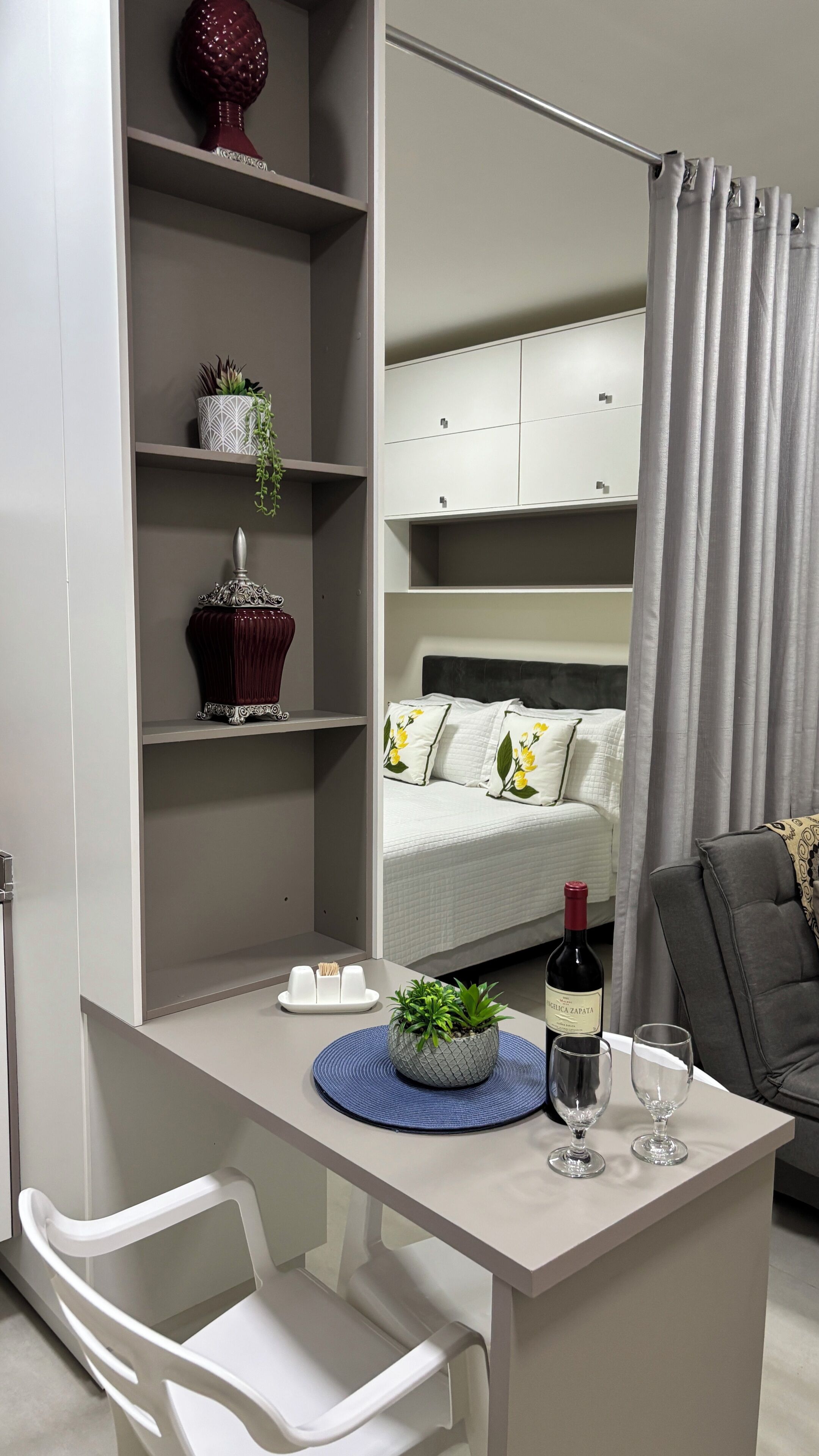 A modern bedroom with a bed, a bedside table with a wine bottle and glasses, a chair, and a desk with a potted plant.