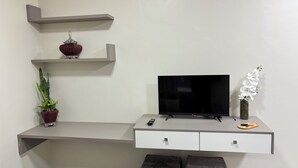 A modern living room with a flat-screen TV, a potted plant, a vase with white flowers, and a remote control on a grey console table. - Ravello Residential AP03 Foz do Iguaçu (Foz do Iguaçu)