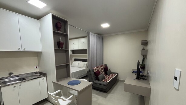 A compact living space with a kitchenette, a dining area with a small table and chair, a sofa, and a TV. - Ravello Residential AP03 Foz do Iguaçu (Foz do Iguaçu)
