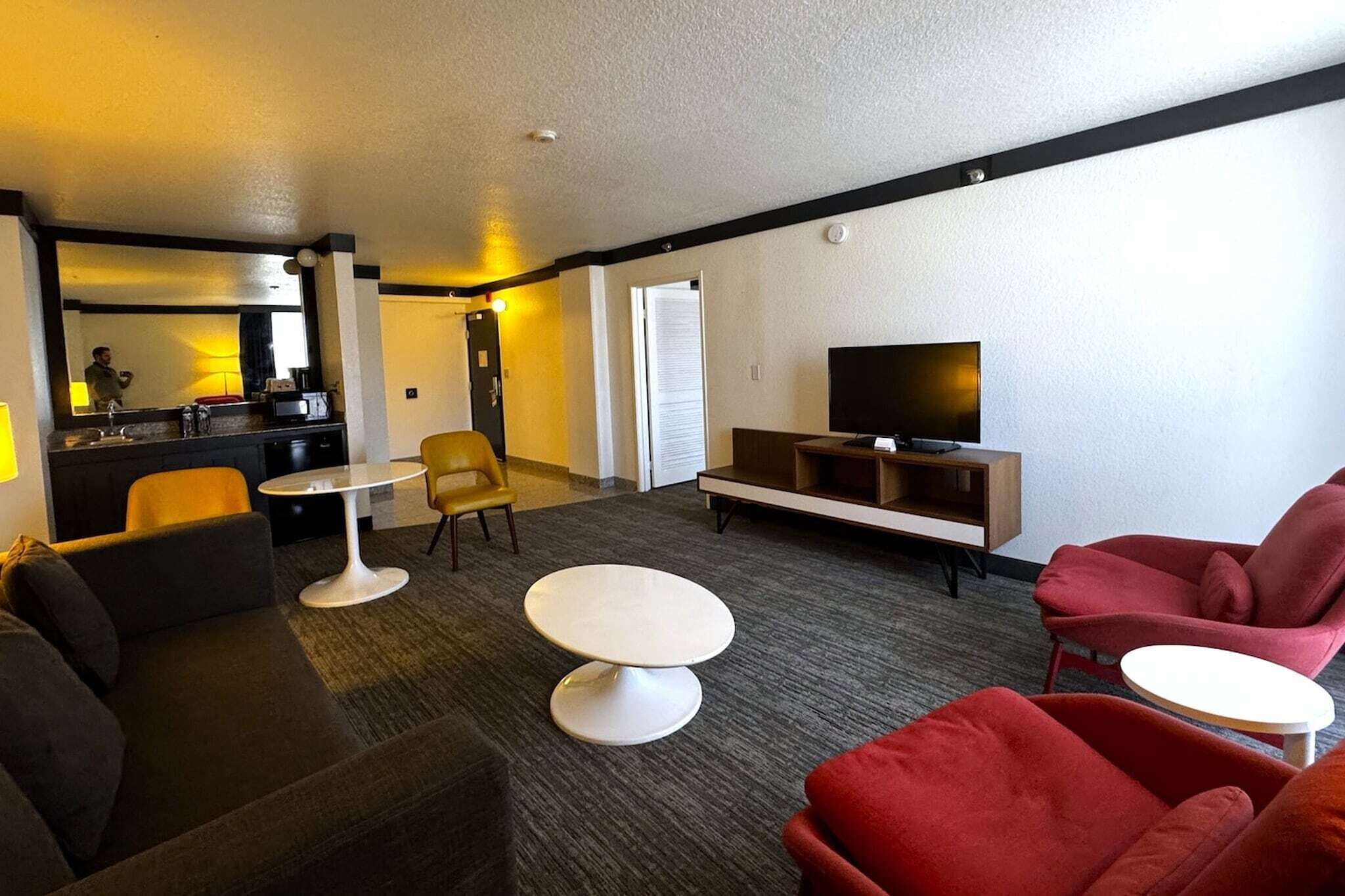 Une chambre d’hôtel avec un canapé, deux fauteuils, une petite table ronde, un téléviseur posé sur un meuble et un miroir.