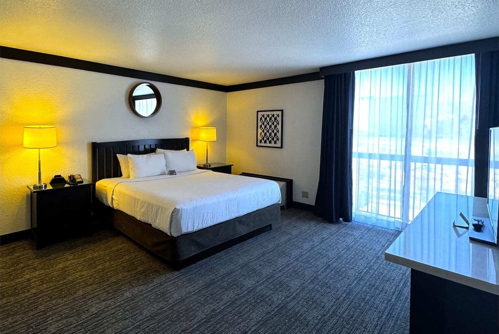 Une chambre d’hôtel avec un lit, deux lampes de chevet, un miroir rond, un tableau encadré, un bureau et une grande fenêtre avec des rideaux.