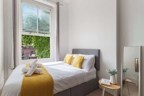 A neatly made bed with pillows, a bedside table with a book, and a window with a view of greenery. - Standard Studio (London)